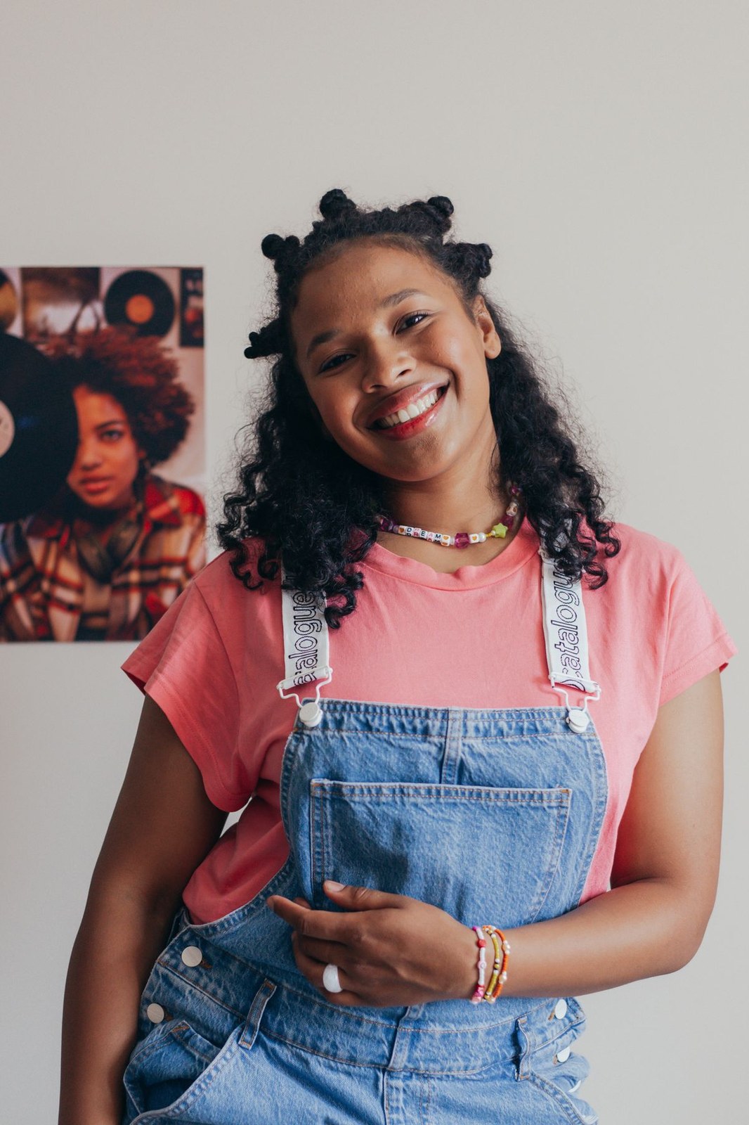 Smiling woman in plus size overalls with curly hair in blue denim dungarees over a pink tee
